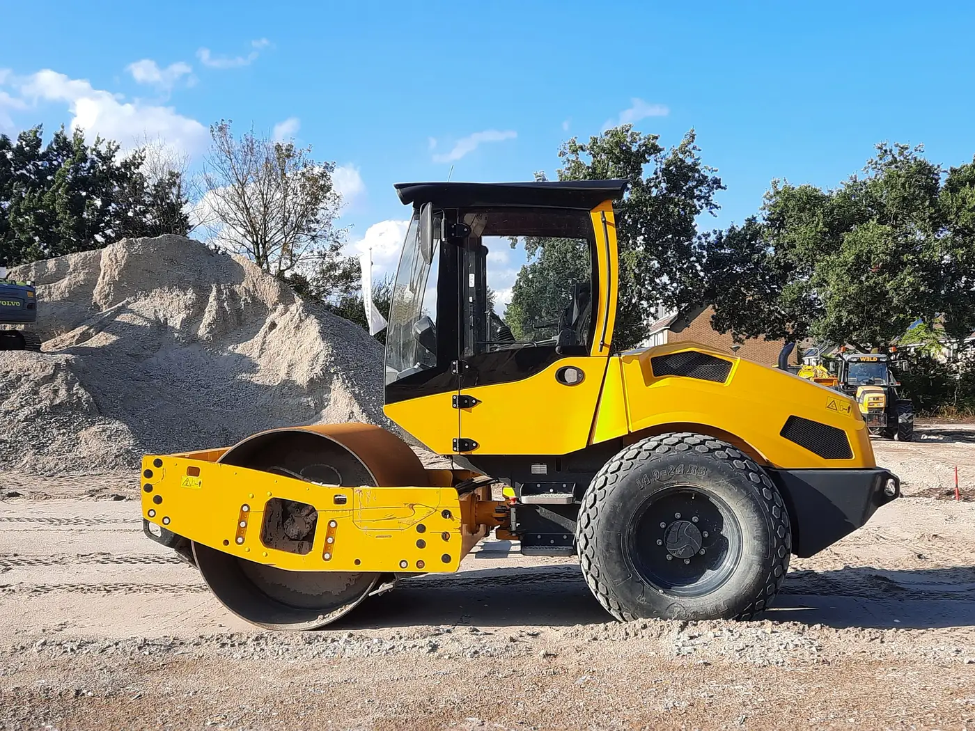 A yellow smooth-drum roller compactor viewed from the side on a dirt construction site, drum and cab visible.